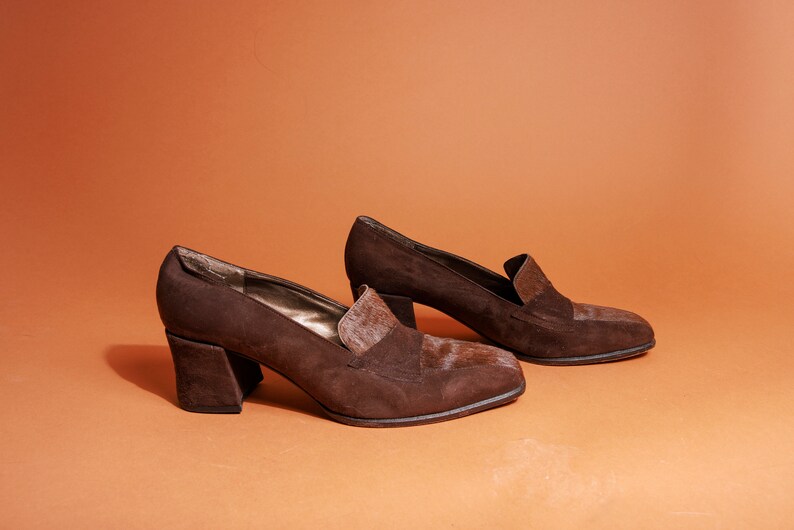 May include: A pair of brown suede heeled shoes with a block heel. The shoes have a pointed toe and a small flap detail on the top.