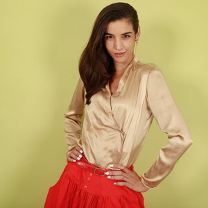May include: A woman wearing a gold satin blouse with a red skirt and a gold belt. She is standing in front of a light green background.