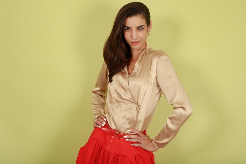 May include: A woman wearing a gold satin blouse with a red skirt and a gold belt. She is standing in front of a light green background.
