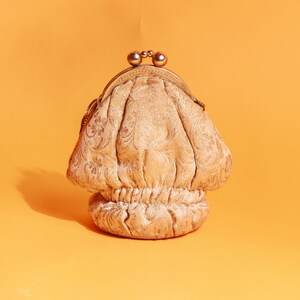 Puede incluir: Un monedero beige de estilo vintage con un estampado floral decorativo. El bolso tiene forma redondeada con un cierre metálico y una base fruncida. El fondo es de color naranja liso.