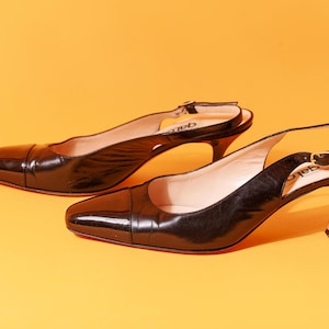May include: A pair of black leather slingback heels with pointed toes. The shoes have a thin heel and a buckle closure on the slingback strap. The heels are set against a yellow background.