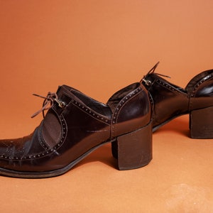 Pu&ograve; includere: Un paio di scarpe con tacco in pelle marrone con un design a lacci e un dettaglio a taglio sul lato. Le scarpe hanno un tacco spesso e una punta a punta.