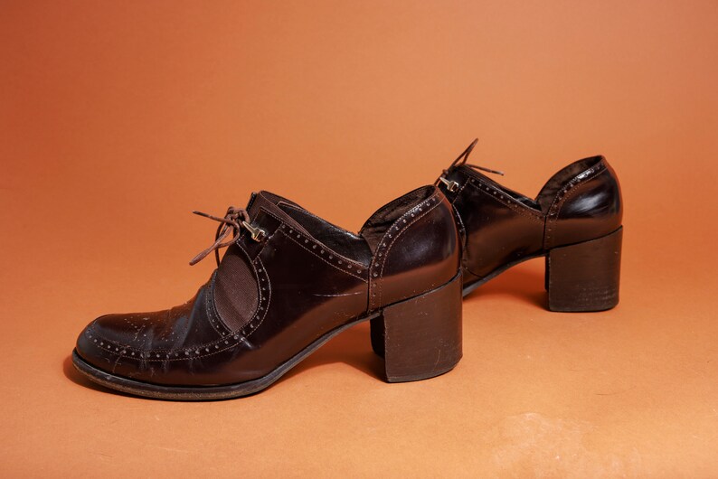 Pu&ograve; includere: Un paio di scarpe con tacco in pelle marrone con un design a lacci e un dettaglio a taglio sul lato. Le scarpe hanno un tacco spesso e una punta a punta.