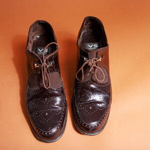 Pu&ograve; includere: Un paio di scarpe marroni in pelle con lacci e design brogue. Le scarpe hanno una fodera in pelle marrone e una suola in pelle nera. Le scarpe hanno un piccolo cartellino metallico sul lato che dice "VS Made in Italy".