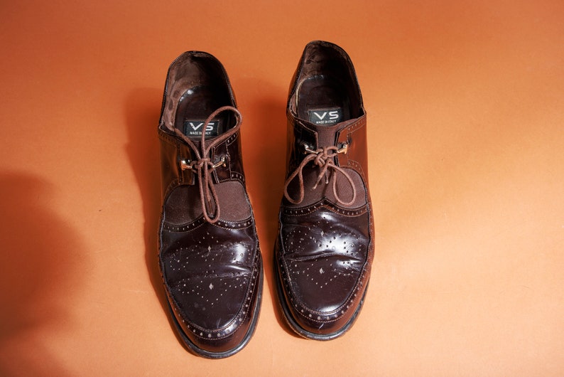 Pu&ograve; includere: Un paio di scarpe marroni in pelle con lacci e design brogue. Le scarpe hanno una fodera in pelle marrone e una suola in pelle nera. Le scarpe hanno un piccolo cartellino metallico sul lato che dice "VS Made in Italy".