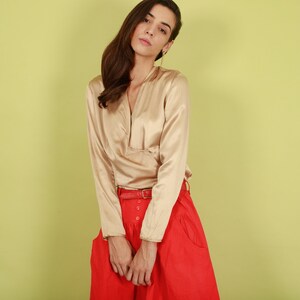 May include: A woman wearing a long-sleeved, beige satin blouse with a V-neck and a red skirt with a belt.