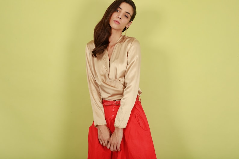 May include: A woman wearing a long-sleeved, beige satin blouse with a V-neck and a red skirt with a belt.