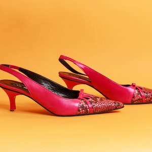 May include: A pair of fuchsia slingback heels with pointed toes and small bows. The toe of each shoe has a snakeskin pattern. The heels are approximately 2 inches high. The shoes are set against an orange background.