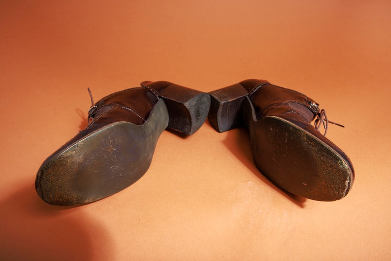 Pu&ograve; includere: Un paio di scarpe marroni in pelle con tacco e lacci. Le scarpe sono mostrate dal basso, con le suole rivolte verso la fotocamera.