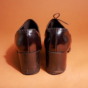 Pu&ograve; includere: Un paio di scarpe con tacco in pelle marrone con punta a punta e design con lacci. Le scarpe hanno un tacco spesso e una finitura lucida.