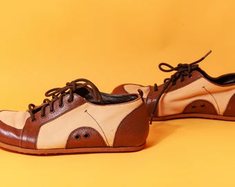 May include: A pair of brown and beige trainers with dark brown laces. The shoes have a rounded toe and a textured brown panel on the heel. The trainers are set against a yellow background.