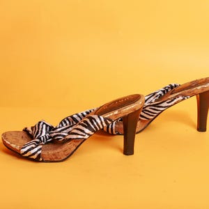 Puede incluir: Un par de sandalias de tacón alto con estampado de cebra. Las sandalias tienen una plantilla de corcho, una tira de tela con estampado de cebra en blanco y negro y un tacón marrón oscuro. Los zapatos están sobre un fondo amarillo.