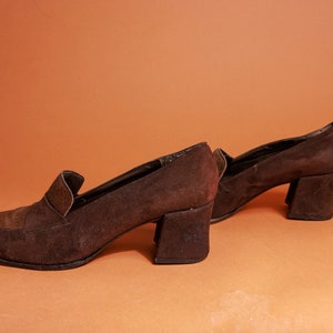 May include: A pair of brown suede heeled shoes with a chunky heel. The shoes have a pointed toe and a slip-on design.