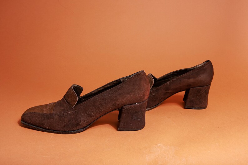 May include: A pair of brown suede heeled shoes with a chunky heel. The shoes have a pointed toe and a slip-on design.