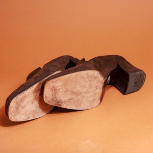 May include: A pair of brown suede heeled shoes with worn soles. The shoes are placed side by side on a brown surface.