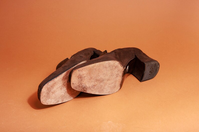 May include: A pair of brown suede heeled shoes with worn soles. The shoes are placed side by side on a brown surface.