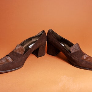 May include: A pair of brown suede heeled shoes with a contrasting brown and white patterned detail on the toe and heel.