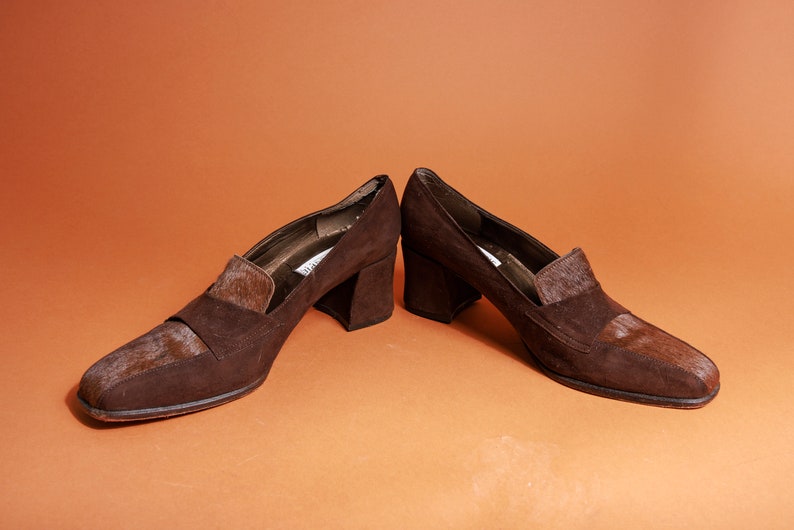 May include: A pair of brown suede heeled shoes with a contrasting brown and white patterned detail on the toe and heel.