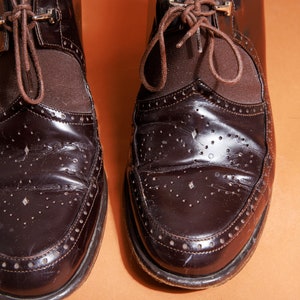 Pu&ograve; includere: Un paio di scarpe in pelle marrone con lacci e un design brogue. Le scarpe hanno una finitura lucida e sono usurate.