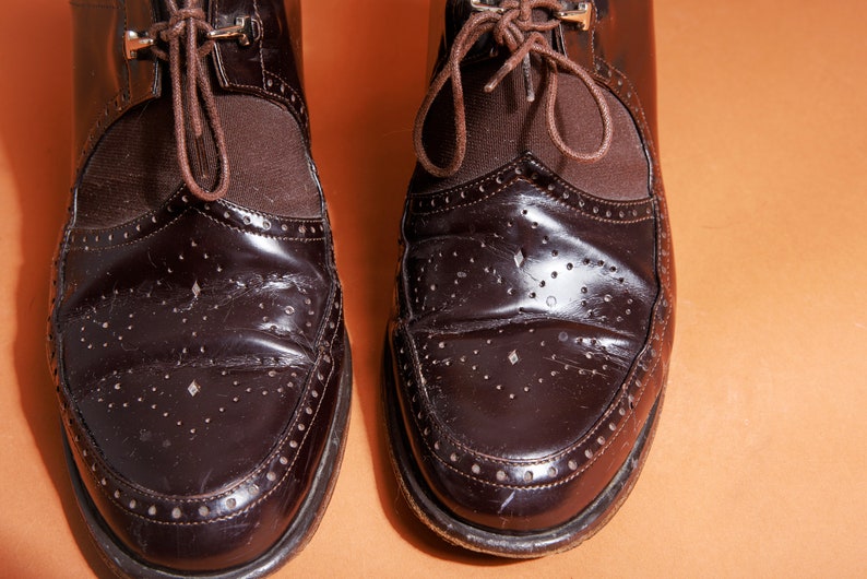 Pu&ograve; includere: Un paio di scarpe in pelle marrone con lacci e un design brogue. Le scarpe hanno una finitura lucida e sono usurate.