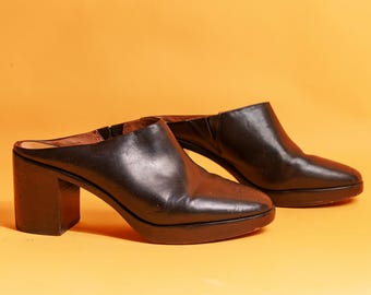 Puede incluir: Un par de mulas de tacón de cuero negro. Los zapatos tienen una punta redondeada y un tacón de bloque. El fondo es de color naranja liso. Los zapatos son de estilo clásico.