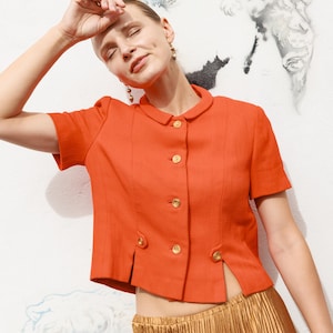 May include: A short-sleeved, bright orange cropped jacket featuring a rounded collar, a textured fabric finish, and five gold-toned circular buttons down the front. The jacket includes two decorative button details at the lower front hem.