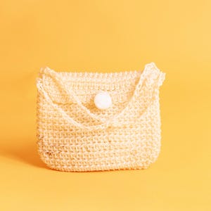 Può includere: Una piccola borsa all'uncinetto color crema, con patta e chiusura a bottone bianco. La borsa ha un aspetto strutturato e un cinturino intrecciato. Lo sfondo è giallo caldo uniforme.