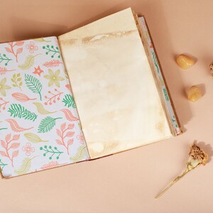 K&ouml;nnte beinhalten: Ein offenes Tagebuch mit einem floralen Muster auf dem Einband und leeren Seiten. Das Tagebuch liegt auf einer hellbraunen Oberfl&auml;che mit zwei getrockneten Blumen und zwei kleinen orangen Steinen.