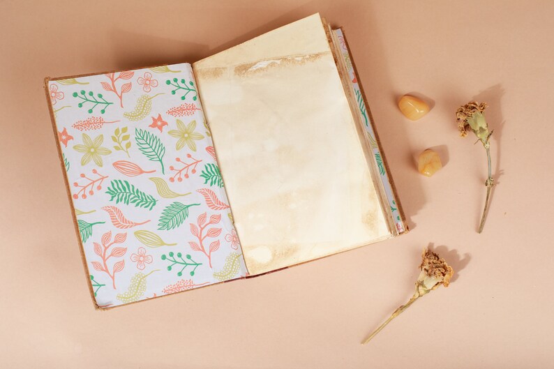 K&ouml;nnte beinhalten: Ein offenes Tagebuch mit einem floralen Muster auf dem Einband und leeren Seiten. Das Tagebuch liegt auf einer hellbraunen Oberfl&auml;che mit zwei getrockneten Blumen und zwei kleinen orangen Steinen.