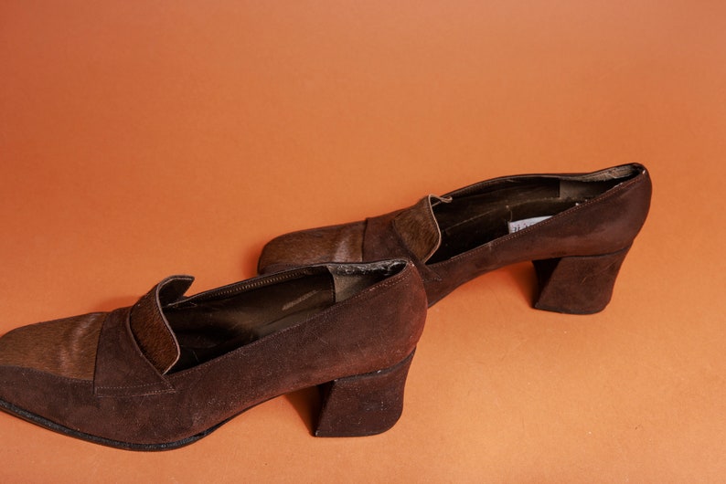 May include: A pair of brown suede heeled shoes with a contrasting brown leather toe cap. The shoes have a chunky block heel.