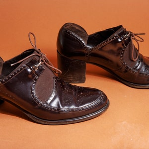 Pu&ograve; includere: Un paio di scarpe marroni in pelle con tacco, lacci sul davanti e un motivo di cuciture decorative. Le scarpe hanno un tacco spesso e una punta a punta.