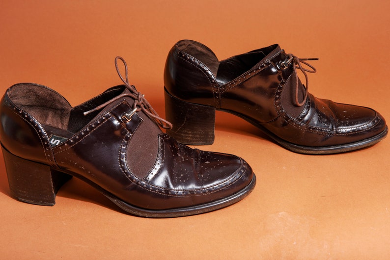 Pu&ograve; includere: Un paio di scarpe marroni in pelle con tacco, lacci sul davanti e un motivo di cuciture decorative. Le scarpe hanno un tacco spesso e una punta a punta.