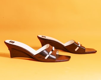 Puede incluir: Un par de sandalias de cuña de cuero marrón con detalles blancos. Los zapatos de punta abierta tienen una punta cuadrada y un tacón de cuña. Las sandalias están sobre un fondo naranja liso.