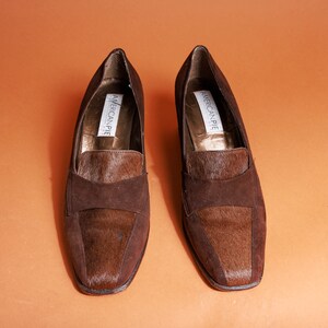 May include: A pair of brown suede loafers with a brown and white fur trim. The loafers have a square toe and a low heel. The brand name "American Pie" is printed on the inside of each shoe.