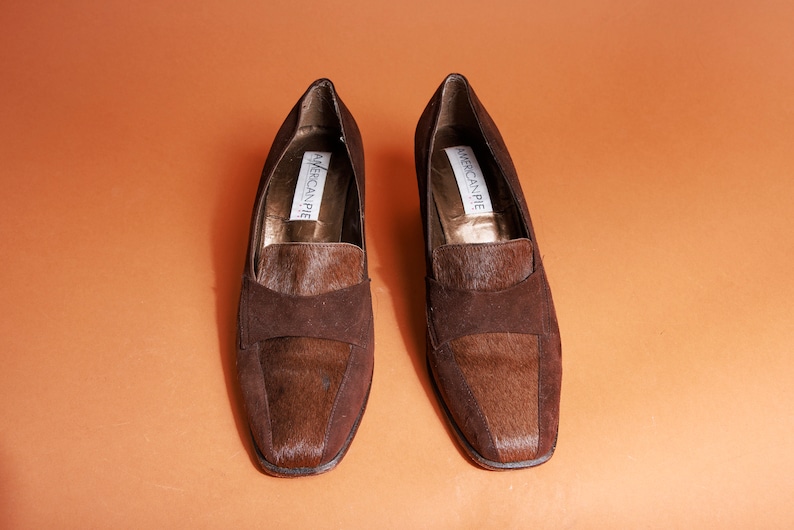 May include: A pair of brown suede loafers with a brown and white fur trim. The loafers have a square toe and a low heel. The brand name "American Pie" is printed on the inside of each shoe.
