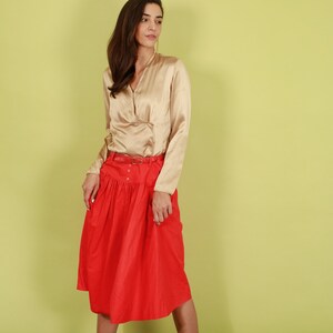 May include: A woman wearing a gold satin blouse with a V-neck and long sleeves, paired with a red midi skirt with a gathered waist and a belt.