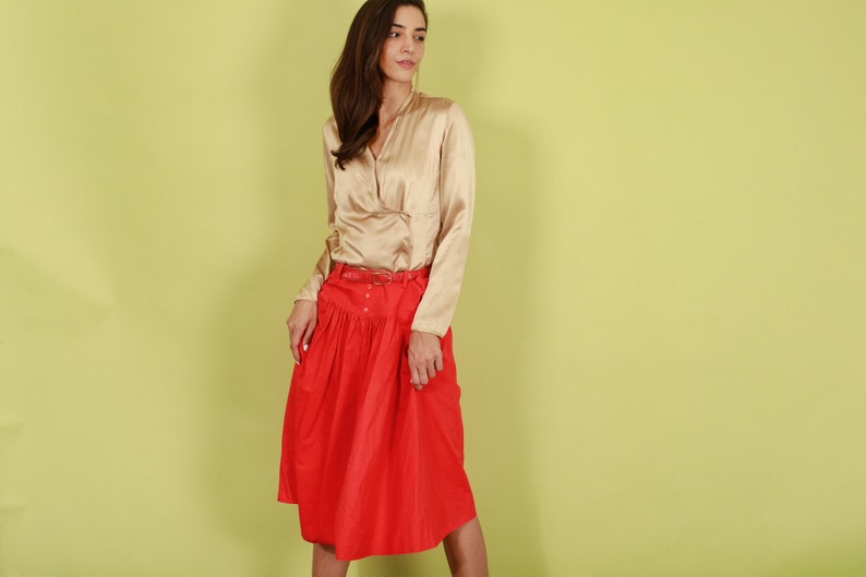 May include: A woman wearing a gold satin blouse with a V-neck and long sleeves, paired with a red midi skirt with a gathered waist and a belt.