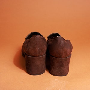 May include: A pair of brown suede heeled shoes with a chunky heel. The shoes are closed-toe and have a simple design.