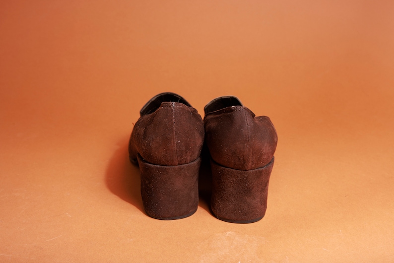 May include: A pair of brown suede heeled shoes with a chunky heel. The shoes are closed-toe and have a simple design.