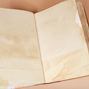 K&ouml;nnte beinhalten: Ein offenes, leeres Tagebuch mit braunen Lederbez&uuml;gen und gealterten, cremefarbenen Seiten. Das Tagebuch ist auf einer leeren Doppelseite ge&ouml;ffnet, wobei die Seiten an den R&auml;ndern leicht gewellt sind. Das Tagebuch liegt auf einer hellbraunen Oberfl&auml;che.
