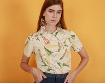 May include: A woman wearing a cream-colored short-sleeved shirt with a floral print and blue jeans. She is wearing large gold earrings.