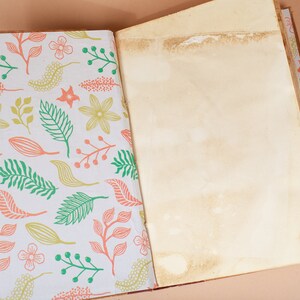 K&ouml;nnte beinhalten: Ein offenes Tagebuch mit einem wei&szlig;en Einband, der ein Blumenmuster in Gr&uuml;n-, Rosa- und Gelbt&ouml;nen zeigt. Die Seiten sind leer und das Tagebuch liegt auf einer hellbraunen Oberfl&auml;che.