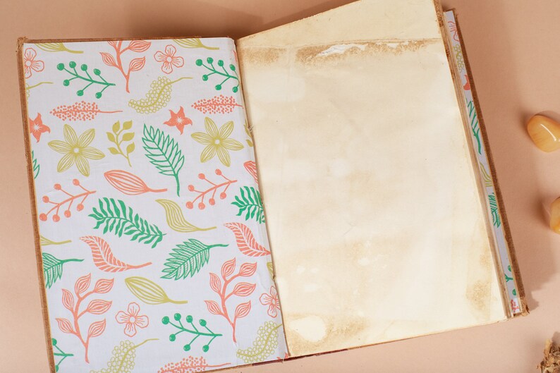 K&ouml;nnte beinhalten: Ein offenes Tagebuch mit einem wei&szlig;en Einband, der ein Blumenmuster in Gr&uuml;n-, Rosa- und Gelbt&ouml;nen zeigt. Die Seiten sind leer und das Tagebuch liegt auf einer hellbraunen Oberfl&auml;che.