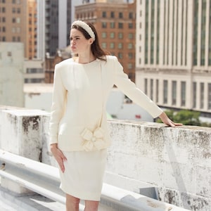 May include: Ivory-colored two-piece suit with a long-sleeved jacket and a knee-length skirt. The jacket features a unique asymmetrical design and a decorative floral accent at the waist. A pearl headband completes the look. The outfit is set against an urban backdrop.