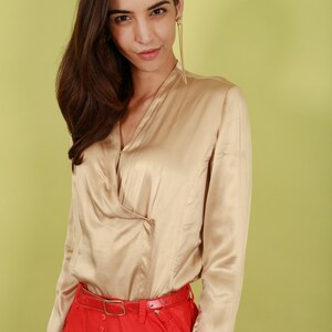 May include: A woman wearing a beige satin blouse with a red skirt and a red belt. The blouse has a V-neck and long sleeves.