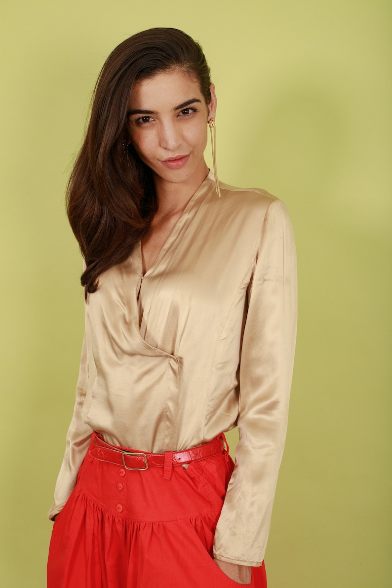 May include: A woman wearing a beige satin blouse with a red skirt and a red belt. The blouse has a V-neck and long sleeves.
