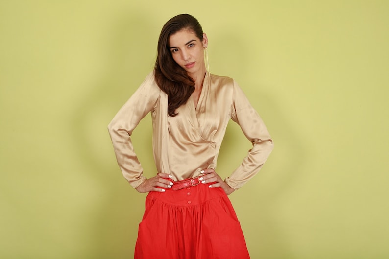 May include: A woman wearing a beige satin blouse with a red skirt and a brown belt. She is standing in front of a light green background.