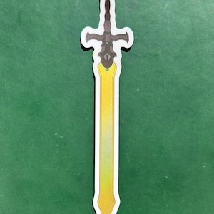 May include: A sticker of a sword with a yellow blade and a dark gray hilt. The blade has a gradient from yellow to white. The hilt has a cross-shaped design with green accents. The sticker is outlined in white.