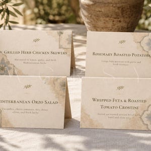 Peut inclure: Quatre étiquettes alimentaires pliées en papier cartonné crème avec du texte gris foncé et des coins décoratifs à motifs de carreaux bleus et dorés. Les cartes présentent des plats comme Grilled Herb Chicken Skewers, Rosemary Roasted Potatoes, Mediterranean Orzo Salad et Whipped Feta & Roasted Tomato Crostini.