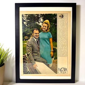 May include: Framed vintage magazine print featuring a smiling couple. The woman wears a turquoise dress and the man a gray suit. The print includes text and a black frame, suitable for wall decor.